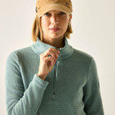 Close-up of a model in an ivy green half-zip fleece top, holding the zip pull, wearing a tan cap.