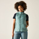 Woman wearing an ivy green quilted gilet over a black T-shirt and blue jeans, one hand in pocket.