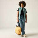 Full-length view of a woman wearing an ivy green quilted gilet, black T-shirt and blue jeans, holding a yellow striped tote bag.