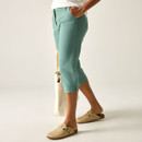 Model wearing green cropped trousers with a white T-shirt, shown from the side with hands in pockets and beige clogs.