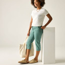Model leaning against a white plinth wearing green cropped trousers and a white T-shirt, holding a cream tote bag and wearing beige clogs.