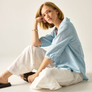 Model seated wearing the light blue pinstripe blouse and white trousers, showing the relaxed fit and sleeve shape.