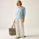 Full-length view of the model in the light blue pinstripe blouse and wide-leg white trousers, holding a striped tote bag.