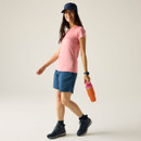 Full-length view of woman wearing a peach marl crew-neck T-shirt, blue shorts and hiking boots, holding an orange water bottle.