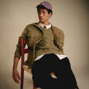 Young man in a casual outfit, wearing a lavender cap and green jacket over a white shirt. He sits relaxed on a chair against a plain backdrop, exuding a laid-back vibe.