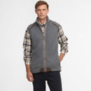 A man wearing a grey vest over a plaid shirt stands against a plain background. He exudes a relaxed, confident demeanour with a casual style.