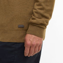 A person wearing a brown jumper with a small logo and dark pants. The focus is on the jumper's texture and the person's hand by their side.
