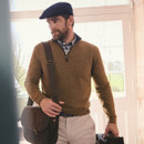 A man wearing a brown jumper and flat cap stands by a window with a serious expression, holding a brown leather satchel and a small bag. Bright, cosy atmosphere.