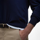 A person in a navy blue jumper with a white vest and beige pants, hand in pocket. The tone is casual and stylish, with a woven belt visible.
