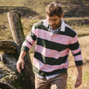 A man walks outdoors wearing a pink and green striped rugby shirt over a plaid shirt. The setting is a rustic field with a wooden fence post, conveying a casual and relaxed mood.