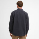 A man with short brown hair facing away, wearing a dark grey button-up shirt and beige pants, stands against a plain white background.