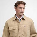 A man wears a khaki jacket with buttoned pockets over a plaid shirt, looking to the side. The expression is neutral. Plain white background.