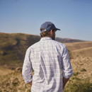 Man in a checkered shirt and blue cap, looking at vast rolling hills under a clear blue sky. The scene conveys solitude and contemplation.