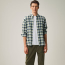 A man stands against a neutral background, wearing a green plaid shirt over a white T-shirt, with olive-green pants. His expression is calm and neutral.