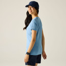 Side view of a woman wearing a light blue outdoor T-shirt with a printed forest design, navy shorts and a navy cap.