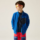 Young boy wearing reflective sunglasses, a blue jacket with black accents, and red shorts with blue pockets. He stands confidently against a neutral background.