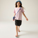 Young girl smiling, wearing a pink T-shirt with red and blue design, black shorts, blue hat, and sandals. She carries a colourful crossbody bag.