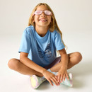 A smiling girl sits cross-legged on a white background, with pink sunglasses, blue butterfly shirt, and colourful sneakers, conveying joy and playfulness.