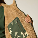 Close-up of a hand holding open a tan, fluffy zip-up jacket, revealing a green inner pocket with a white horse pattern, against a plain background.