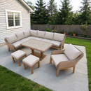 Outdoor patio with light gray sectional sofa, armchair, and two stools surrounding a square table. Set on a concrete slab by a house and lawn, framed by a wooden fence with greenery beyond.