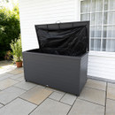 A large, open outdoor storage box with a black interior stands on a tiled patio. It's situated next to a white house with a window, surrounded by greenery.