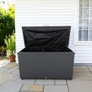 Grey outdoor storage box with an open lid revealing a black interior, placed against a white house with a window. Nearby, a potted plant adds greenery.