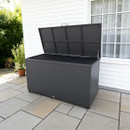 A large black outdoor storage box with an open lid sits on a tiled patio. It's placed against a white house exterior near a potted plant and window.