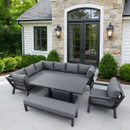 Modern patio with grey sectional sofa, chair, and bench around a table on a stone terrace. Lush greenery and soft lighting create a serene atmosphere.