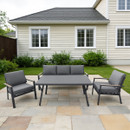 Outdoor patio with grey seating set, including a sofa and two chairs around a table on stone tiles. Cream house and green bushes in background.