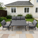 Outdoor patio set with a large grey table, a cushioned sofa, and two matching armchairs on a stone-tiled yard. A beige house and green shrubs are in the background.