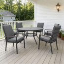 A cosy patio setting with a round grey table and five cushioned chairs on a wooden deck. Green trees and a small house are visible in the background.