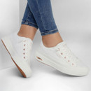Person wearing white trainers with rose gold eyelets, paired with rolled-up blue jeans. The trainers have a platform sole and convey a casual, stylish vibe.
