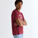 Side view of model wearing a ruby wine Wrangler graphic T-shirt with arms crossed and light blue jeans.