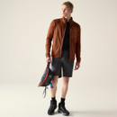 Full-body shot of model in rust-brown midlayer, dark shorts and hiking boots, holding a small sling bag in a studio setting.