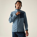 Person stands facing camera, wearing a light blue marl zip-up midlayer with a navy chest panel, hand holding the collar zip.