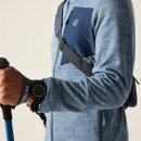 Close-up of the jacket’s navy chest panel with a crossbody strap across it; person holds trekking poles and wears a digital watch.