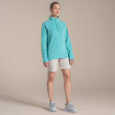 Full-body studio shot of woman in turquoise half-zip fleece and light shorts with trainers.