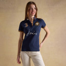 Model wearing a navy Joules Polo Club women’s polo shirt with tan trim, large “3” applique and embroidered crest, paired with cream trousers.