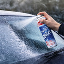 A hand sprays de-icer from a can onto a frosty car windshield, partially clearing the ice. The scene conveys a cold, wintery morning.