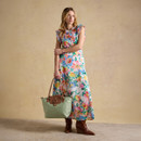 Full-length model in a floral maxi dress holding a sage green Joules tote bag with tan leather handles.