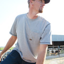 Outdoor photo of a worker wearing a heather gray pocket T-shirt and jeans at a farm, with fencing and a barn in the background.