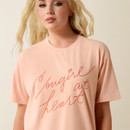 Blonde model wearing a peach short-sleeve T-shirt with cursive “Cowgirl at Heart” lettering across the chest, shown from the waist up on a neutral background.