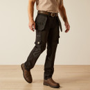 Three-quarter side view of the model stepping forward in black utility pants with large tool pouches and thigh pockets, paired with a tan T-shirt, belt, and brown boots.
