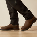 Close-up of brown work boots and the lower legs of black utility pants, showing reinforced seams and small reflective details near the calf.