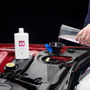 Autoglym Ultimate Screenwash bottle sits in an open engine bay while washer fluid is poured into the reservoir through a funnel.