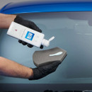 Gloved hands squeeze Autoglym Car Glass Polish onto a foam applicator pad in front of a car windscreen.