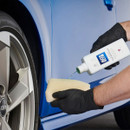 Gloved hands pour Autoglym Intensive Tar Remover onto a cloth in front of a blue car wheel arch for application.