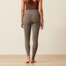 A woman stands barefoot on a light wood floor, wearing a brown tank top and high-waisted grey leggings. She faces away, showing a simple, calm pose.