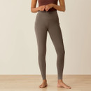 Woman in a brown sleeveless top and taupe high-waisted leggings stands barefoot on light wood floor. Neutral-toned, relaxed setting.