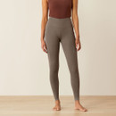 Woman in brown sleeveless top and taupe leggings stands barefoot on a light wooden floor. The setting is minimalistic, conveying a calm, neutral tone.